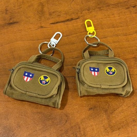 Two small olive drab canvas keychain pouches with top handles and metal clips, placed on a wooden surface. Each pouch has embroidered insignias: a U.