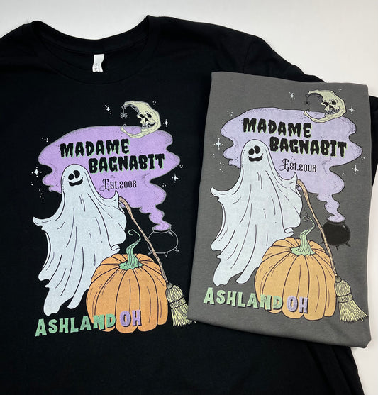 Two black t-shirts, one with a design featuring a white ghost, a pumpkin, and a broom, and the other with a design featuring a gray ghost, a pumpkin, and a broom. Both shirts have text related to the brand 'Madame Bagbabit' and are laid out on a white background.