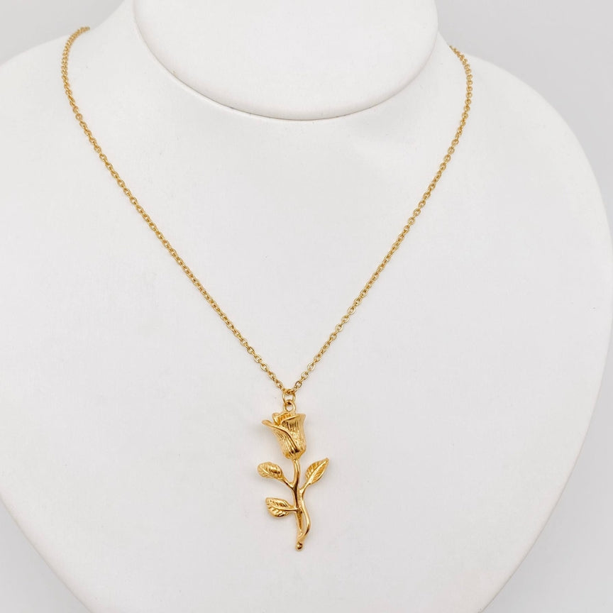 The 18K gold plated Rose pendant necklace by faire is a delicate piece featuring a pendant shaped like a blooming rose with leaves. It is crafted with vacuum electroplating and displayed on a white stand, showcasing its elegant design and intricate details.