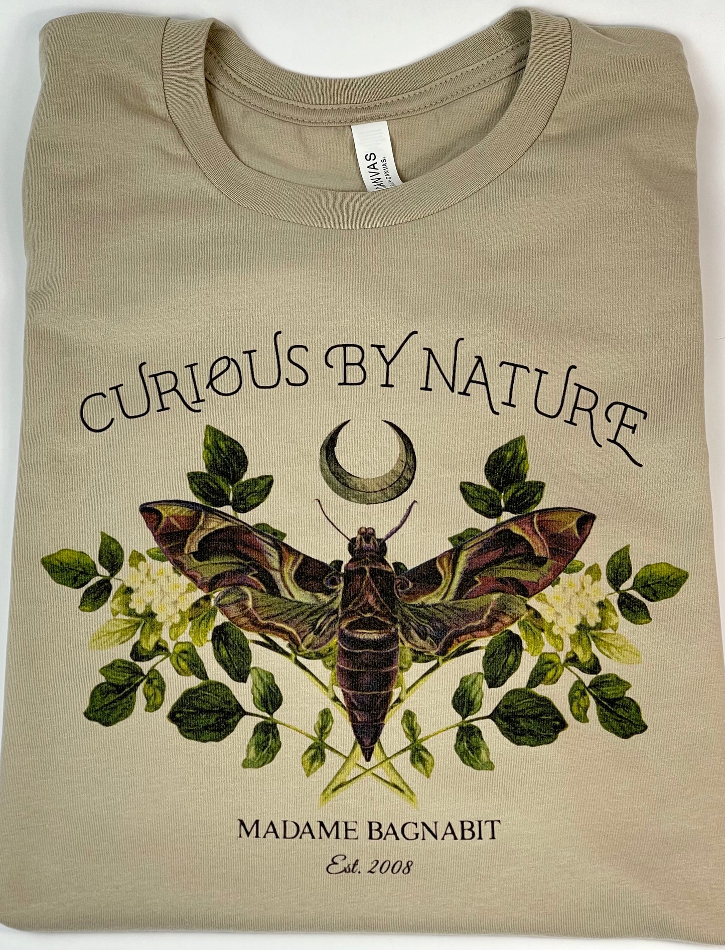 Beige t-shirt with moth and floral design, text 'Curious by Nature' and 'Madame Bagnavit' on a white background