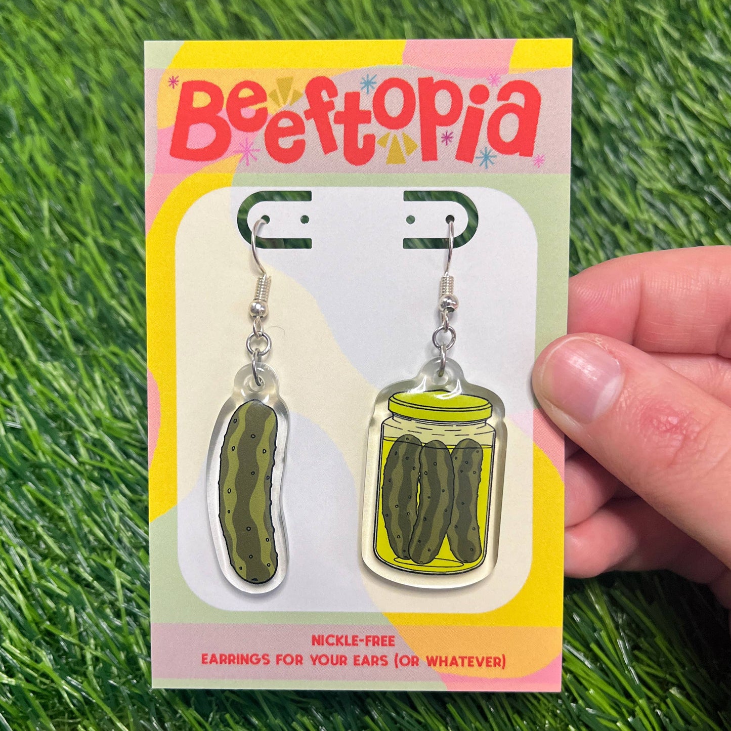 Pickle-themed earrings on a 'Beeftopia' branded card held over green grass.