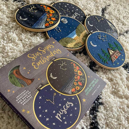 Embroidery hoops with star sign designs and a book titled 'Star Signs Embroidery' on a textured surface.