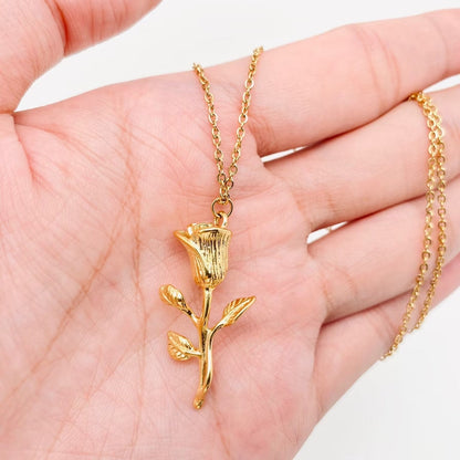 A hand holds the breathtaking 18K gold plated Rose pendant necklace by faire, expertly crafted from stainless steel. The pendant, shaped like a blooming rose with leaves, is elegantly attached to a delicate chain and enhanced through vacuum electroplating for lasting beauty.