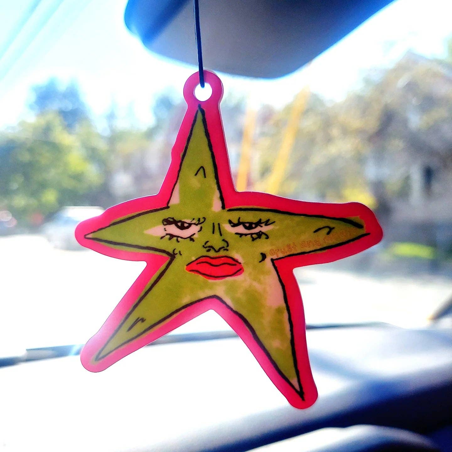 Star-shaped air freshener with facial features hanging from a car's rearview mirror.