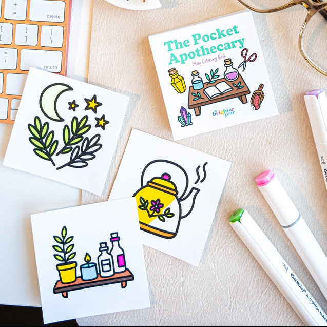 A styled flat-lay of four square white coloring sheets with colorful illustrated designs, including a crescent moon with stars, botanical leaves, a yellow teapot with flowers, and a small apothecary table. The sheets are arranged on a light beige surface with a keyboard, eyeglasses, a crescent moon dish, and marker pens nearby.