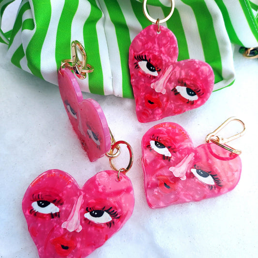Pink heart-shaped keychains with face designs on a white surface with a green and white striped fabric in the background.