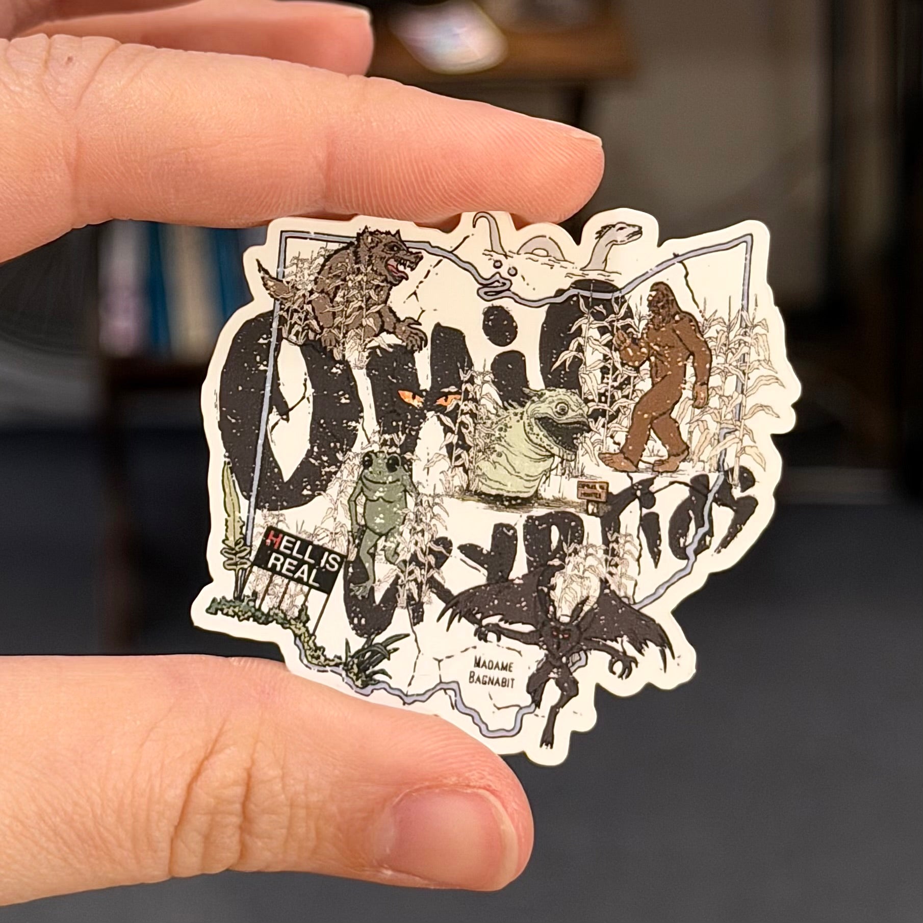 Sticker with various illustrations and text held between two fingers against a blurred background