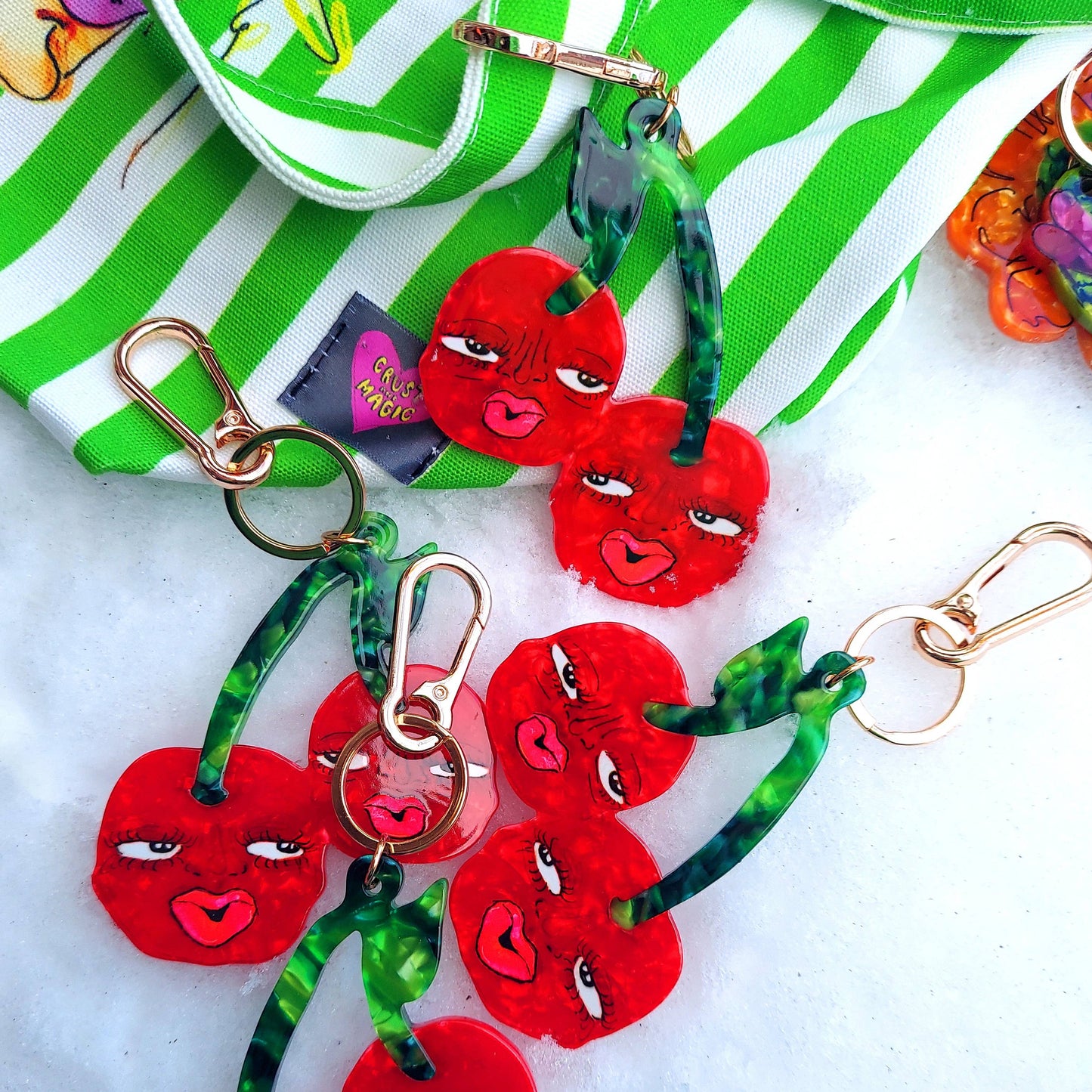 Red cherry keychains with green ribbons on a white surface with a green and white striped fabric in the background.