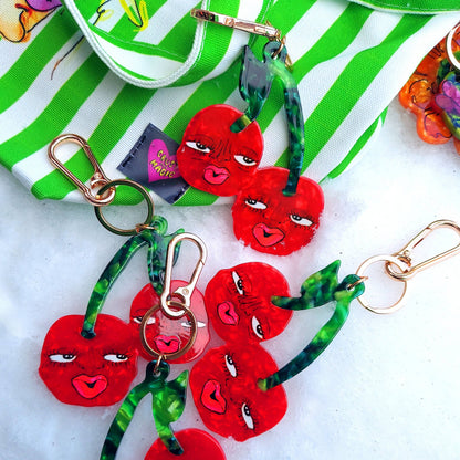 Red cherry keychains with green ribbons on a white surface with a green and white striped fabric in the background.