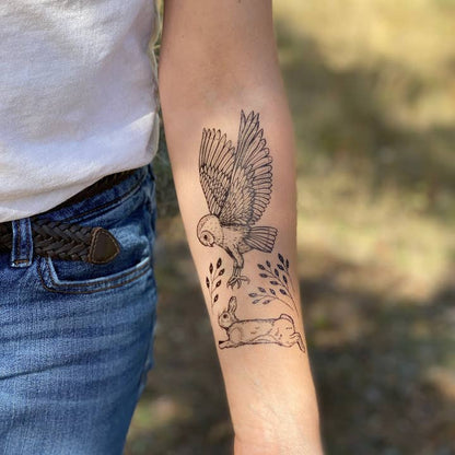 Person wearing a white shirt and blue jeans with a tattoo of an owl and rabbit on their arm.