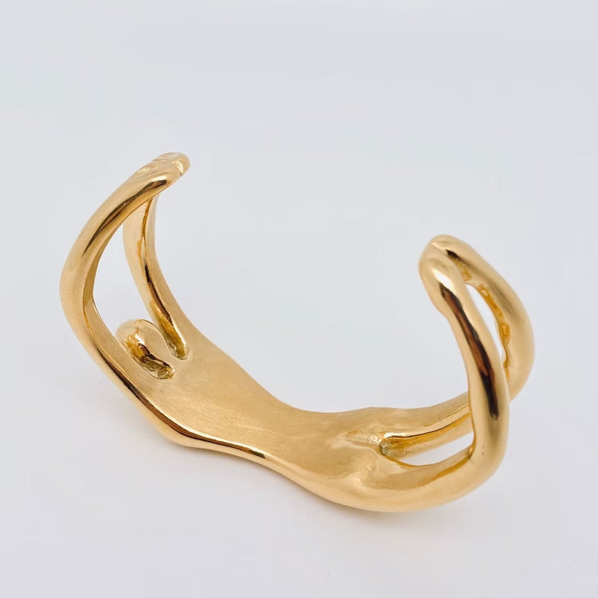 Introducing the "18K gold plated Woman bangle bracelet" by faire: a sleek, abstract cuff crafted from 18K gold-plated stainless steel. This elegant piece is defined by its smooth, flowing curves and open spaces, set against a subtle plain white background.