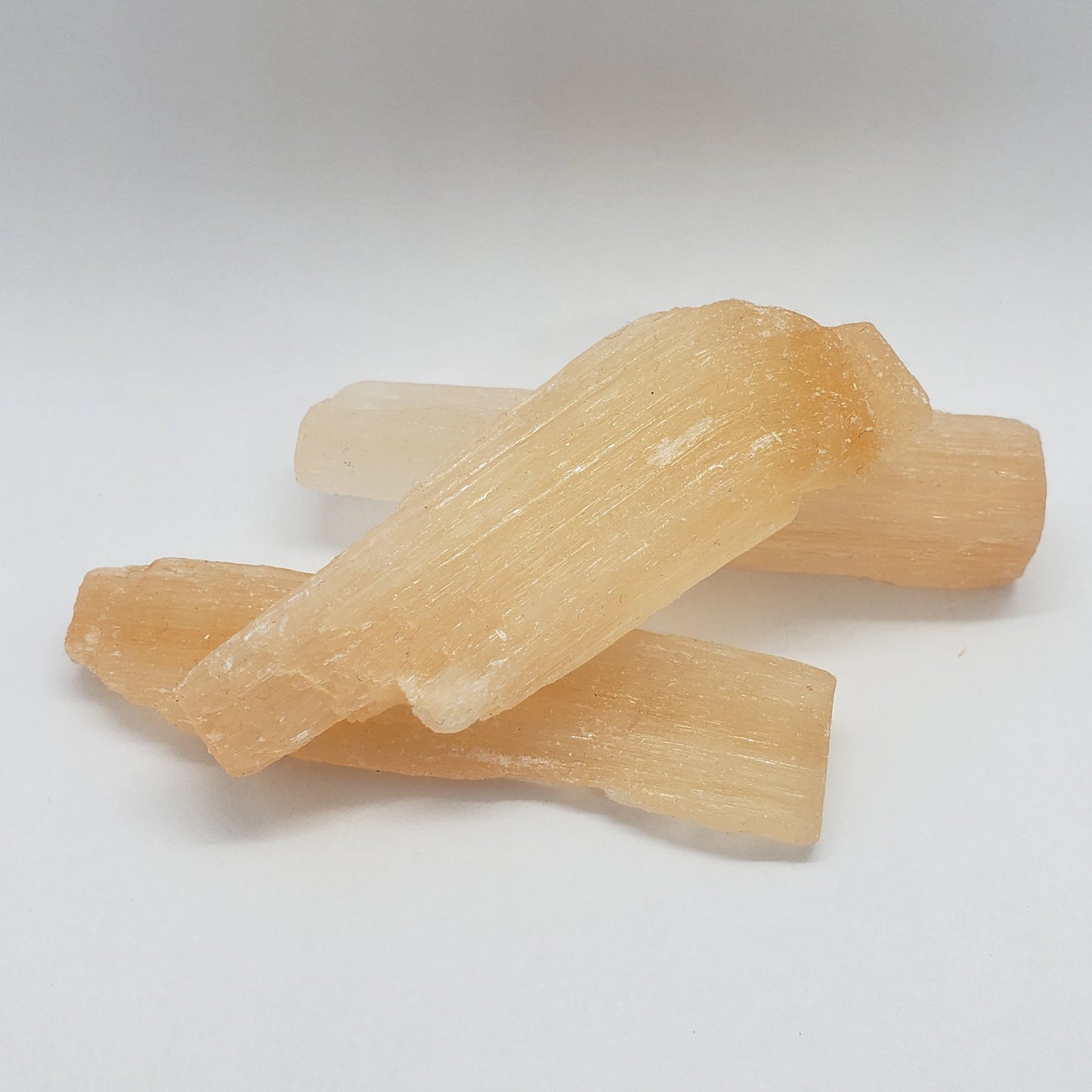 Three translucent, orange selenite sticks from Madame Bagnabit are arranged on a plain white background, showcasing their long, fibrous structure and natural texture.