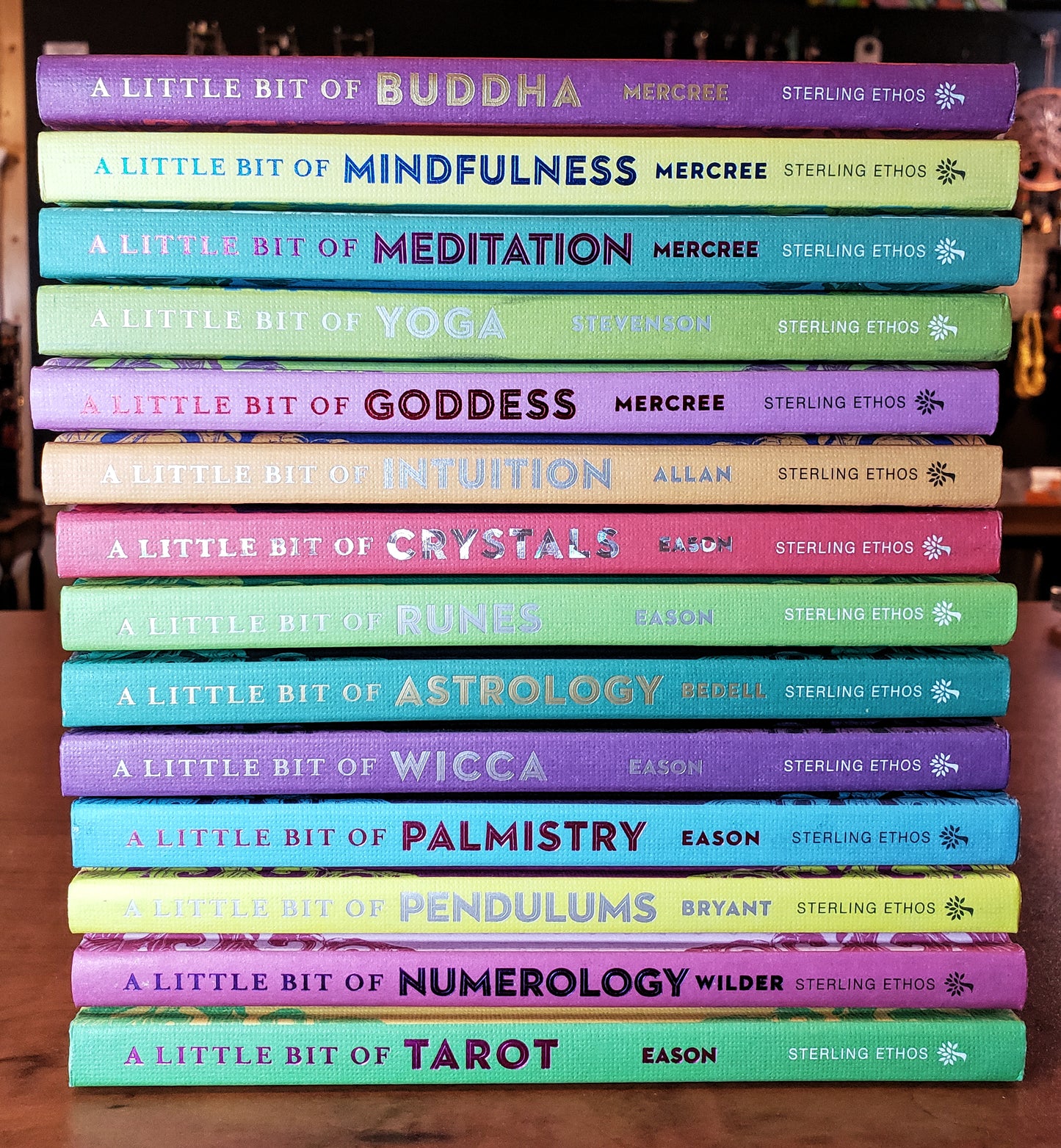 A neatly arranged stack of colorful books from the "A Little Bit of..." series by Sterling features diverse topics such as Buddha, Mindfulness, Meditation, Yoga, and more. Each book stands out with its bright and distinct cover on a wooden surface.