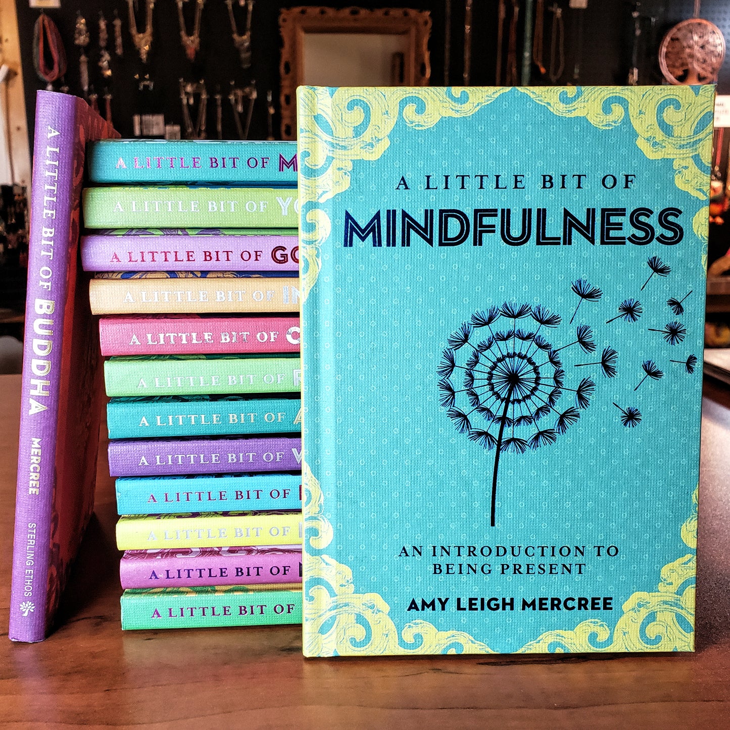 A collection of Sterling's vibrant books titled "A Little Bit of..." featuring themes such as Buddha and Fairies is showcased. Among them, "A Little Bit of Mindfulness" by Amy Leigh Mercree stands upright, adorned with a dandelion on its cover. All the books are displayed on a wooden table.