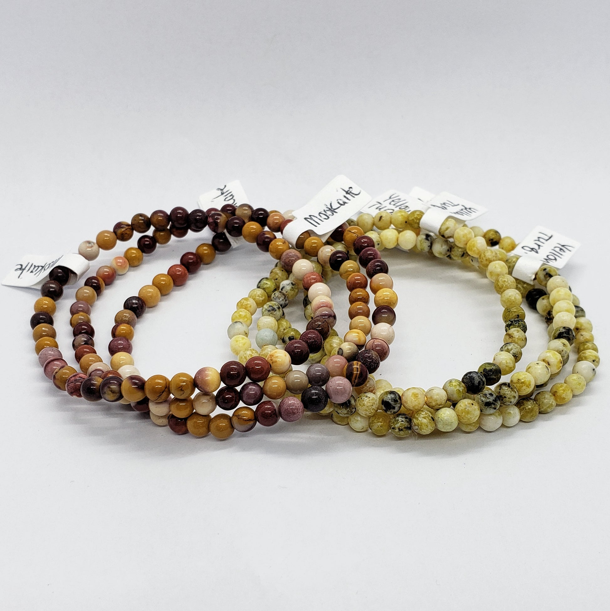 Three sets of Madame Bagnabit's 4mm Energy Stone Bracelets are displayed on a white surface. Each stretch bracelet set showcases beads in earthy tones, featuring shades of brown, beige, and cream. Small tags with the product names are attached to each bracelet set.