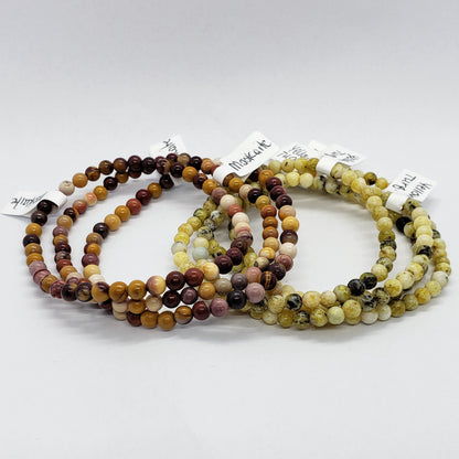 Three sets of Madame Bagnabit's 4mm Energy Stone Bracelets are displayed on a white surface. Each stretch bracelet set showcases beads in earthy tones, featuring shades of brown, beige, and cream. Small tags with the product names are attached to each bracelet set.