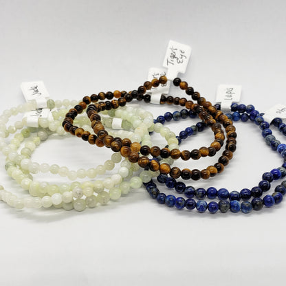 The 4mm Energy Stone Bracelets by Madame Bagnabit are displayed on a white background. Crafted from an assortment of natural stones, including 4mm beads of white jade, tiger's eye, and lapis lazuli, each bracelet comes with a small label attached.